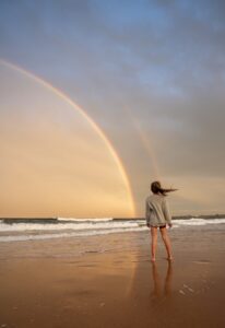 A barefoot woman on a sandy beach gazes at a bright double rainbow over calm ocean waves at sunset.