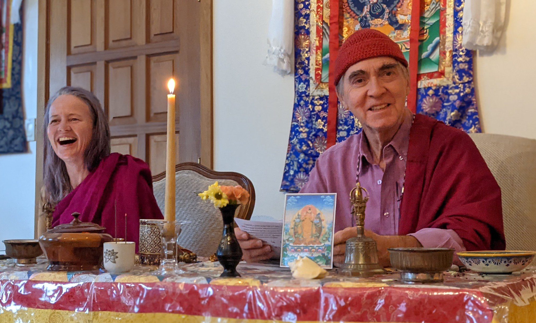 Two people in vibrant traditional robes smile at a table with a candle and flowers in a joyful setting.