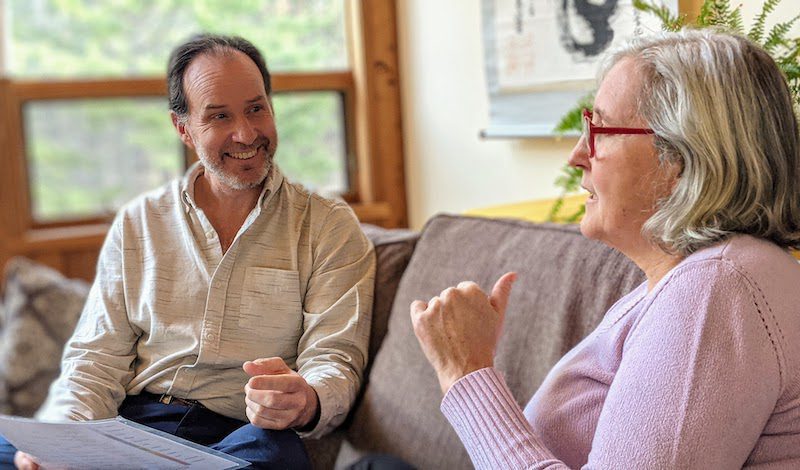 A man with papers and a woman with glasses in a warm conversation on a couch, gesturing during a collaborative meeting.